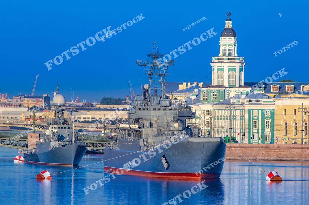 На фотографии изображены военные корабли, стоящие на Неве у Университетской набережной. На заднем плане видна Кунсткамера — одно из самых известных зданий Санкт-Петербурга, символ эпохи Петра Великого. Яркий солнечный день подчёркивает гармонию современной военно-морской силы и классической архитектуры города.